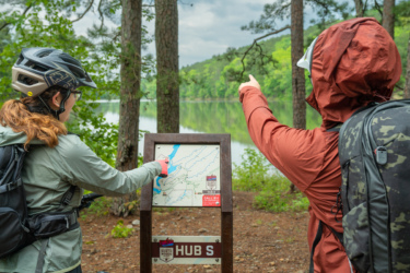 Two people examine a trail map at a scenic outdoor location. One person, wearing glasses and a helmet, points at the map while the other gestures toward the landscape. Tall trees and a body of water are visible in the background, showcasing a lush, green environment. The map indicates a location marked "HUB S." Northwoods Trail System mountain bike trail.