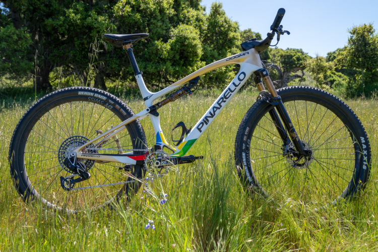A high-performance Pinarello Dogma mountain bike with a white and gold frame, featuring the brand name "Pinarello" prominently displayed. The bike is positioned on lush green grass, surrounded by wildflowers and trees in the background. The tires are designed for rough terrain, and the bike showcases various high-end components, including a visible rear derailleur and a suspension system.