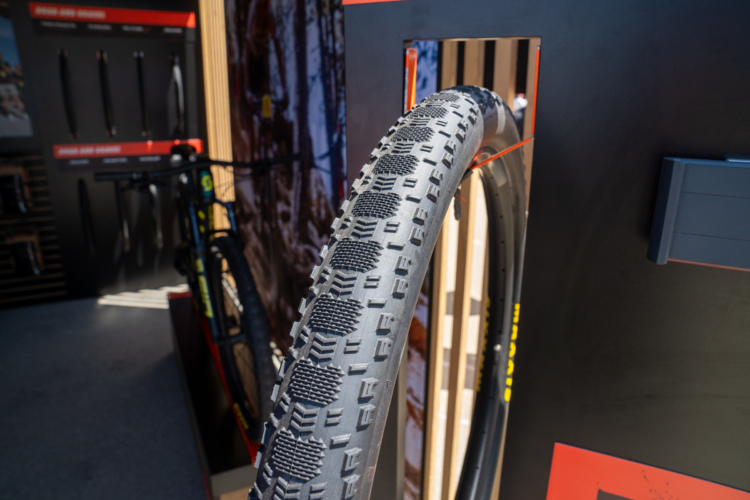 A close-up image of a mountain bike tire displayed in a retail setting. The tire features a textured tread pattern designed for off-road traction, and is partially positioned within a display showcasing bike accessories in the background. Bright lighting highlights the details of the tire against a backdrop of promotional materials.