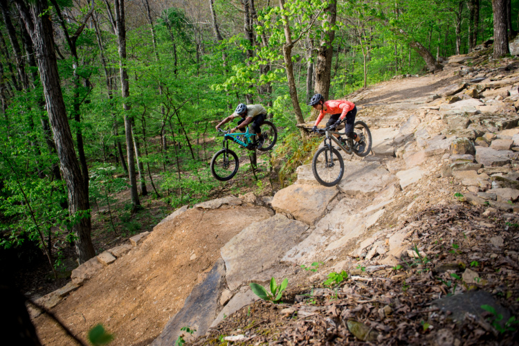Two mountain bikers jumping off a rocky ramp on a wooded trail surrounded by green trees and foliage.