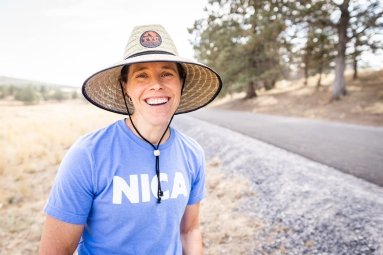 A smiling person wearing a large straw hat and a blue t-shirt stands outdoors near a gravel path, with trees and a dry landscape in the background.