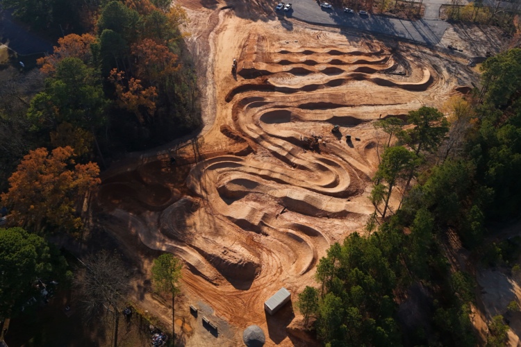 Aerial view of a dirt bike track under construction, featuring winding paths and jumps, surrounded by green trees and fall foliage. Nearby, parked vehicles and a storage container are visible.