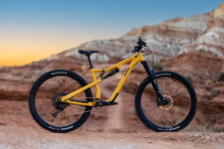 A yellow mountain bike positioned on a dirt path with rocky terrain, set against a backdrop of colorful canyon formations and a sunset sky. The bike features wide black tires with "Maxxis" branding and a sturdy frame, highlighting its design for off-road riding.