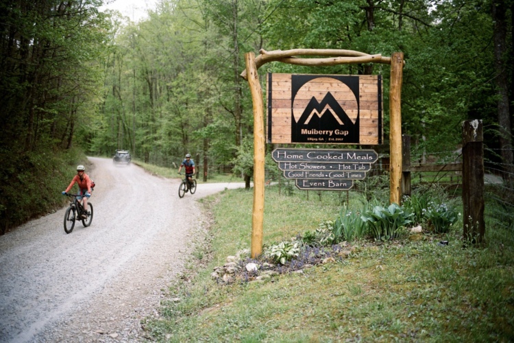 A gravel road winding through a lush green forest, featuring a wooden sign for Mulberry Gap that highlights services like home-cooked meals, hot showers, and an event barn. Two cyclists ride along the road, with a vehicle in the background. Nearby flowers are visible at the base of the sign.