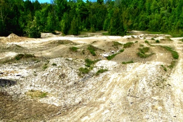 Image of a dirt biking trail in a sunny outdoor setting, featuring winding paths of packed earth and gravel surrounded by lush green trees and a clear blue sky. Harmony Valley Conservation Area mountain bike trail.