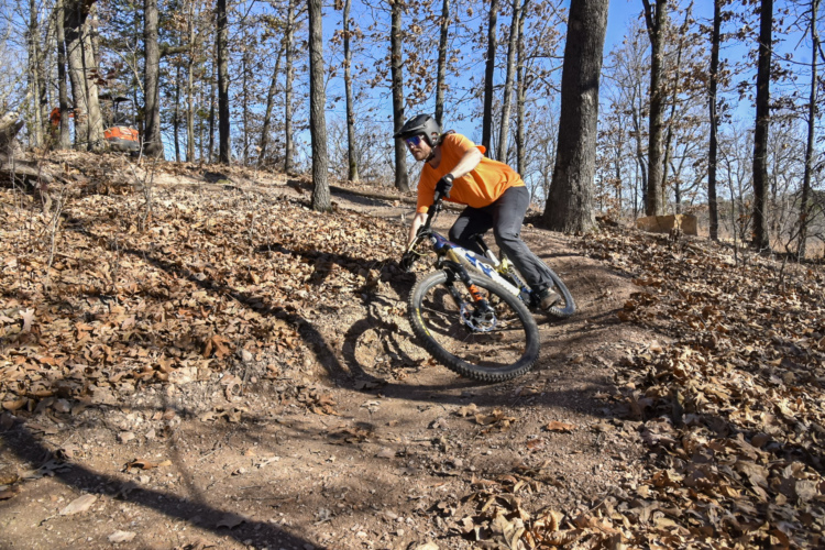 A mountain biker in an orange shirt navigates a winding dirt trail through a wooded area, surrounded by trees and fallen leaves on the ground. The biker leans into the turn for stability, showcasing the energetic movement typical of mountain biking. The clear blue sky peeks through the branches, adding to the outdoor atmosphere.