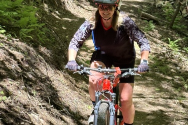 A person riding a mountain bike on a forest trail, smiling while wearing a helmet and cycling attire. Sunlight filters through the trees, illuminating the path. Butter Gap / Trail #123 mountain bike trail.