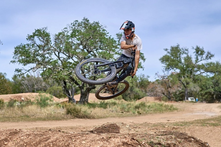 A person performing a stunt on a mountain bike, airborne over a dirt jump, with trees and a clear blue sky in the background.