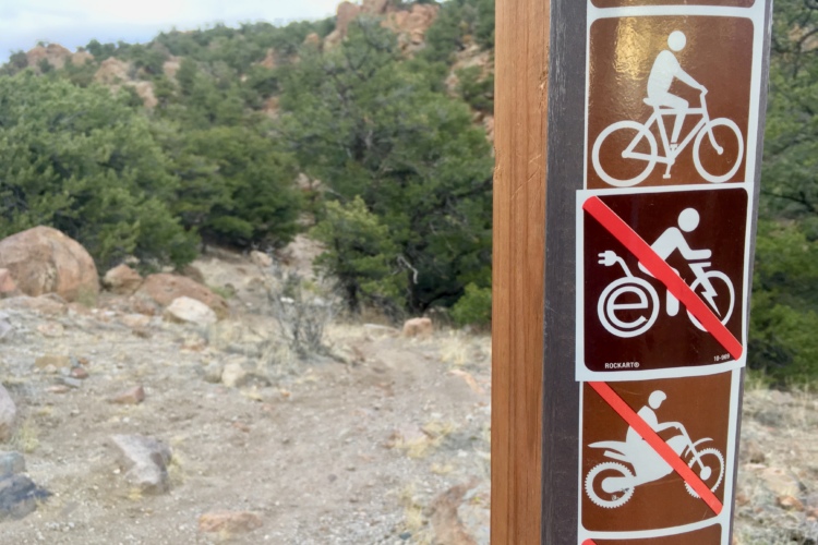A wooden trail sign with various symbols indicating biking rules. The top symbol shows a bicycle, indicating it's allowed, while the second symbol shows an electric bicycle with a red "no" slash across it, indicating it's not permitted. The lower symbols depict a motorcycle and an off-road vehicle, both also marked with red slashes, indicating they are not allowed on the trail. The background features a rocky terrain with green trees.
