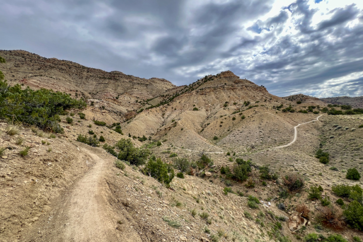 A winding dirt path leads through rugged, hilly terrain under a cloudy sky. The landscape features rolling hills with sparse vegetation and rocky outcrops, creating a dramatic and natural scene.