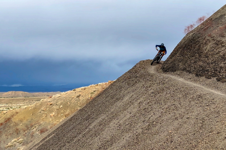 A mountain biker navigates a winding dirt path along a steep, rocky hillside under an overcast sky, with distant hills visible in the background.