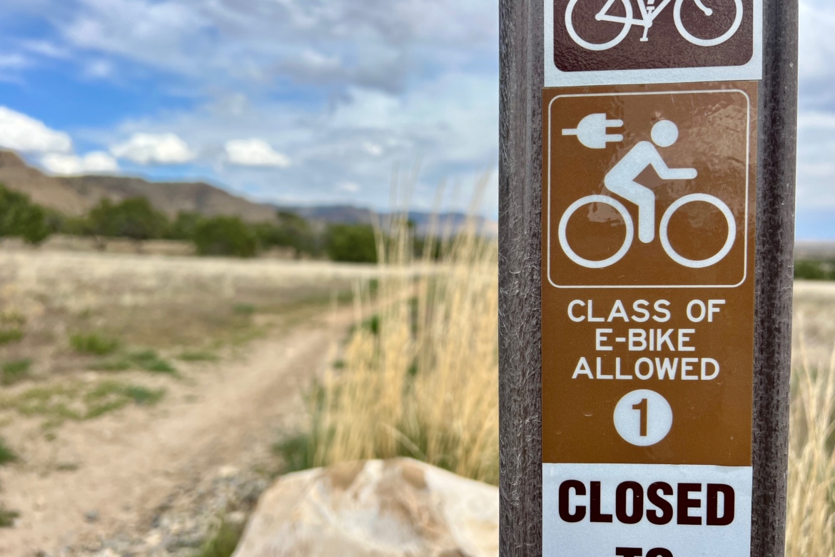 A close-up of a trail sign indicating that class 1 e-bikes are allowed, with a bike icon and an electric plug symbol. The sign also displays a "closed" label at the bottom. In the background, a natural landscape with grassy areas and distant mountains under a partly cloudy sky is visible.