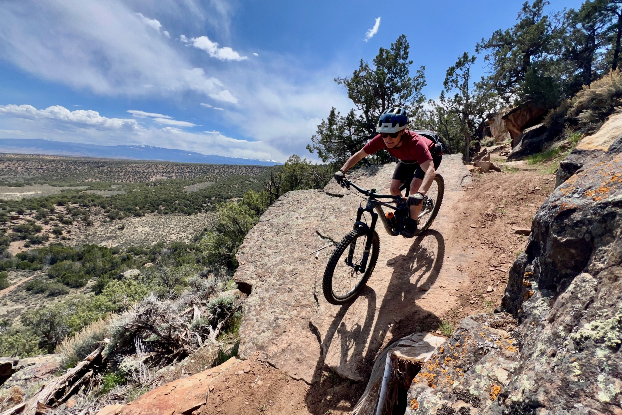 A mountain biker navigates a rocky trail on a sunny day, with a mountainous landscape visible in the background. The biker wears a helmet and rides a black bike, skillfully maneuvering around a large rock. Lush greenery surrounds the trail, creating a picturesque outdoor setting. Electric Hills mountain bike trail.