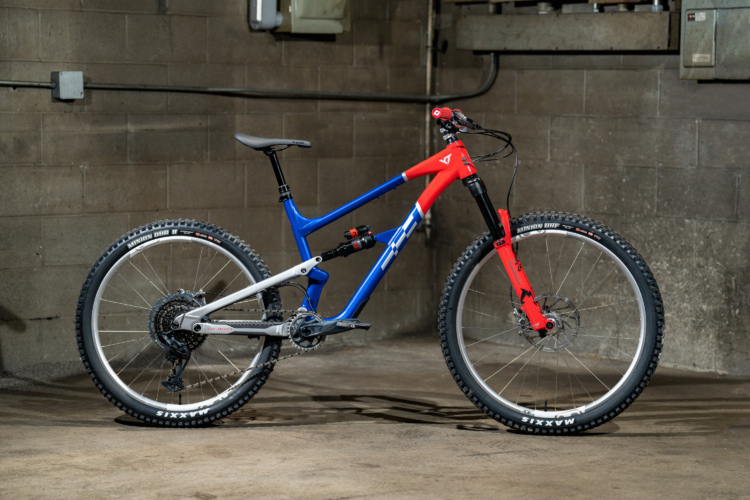 Alt tag: A mountain bike with a blue and red frame, equipped with thick tires and suspension, positioned in a garage environment with a gray concrete wall in the background.