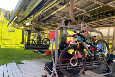 Two mountain bikes are suspended on a ski lift as it prepares for an uphill journey, with people seated in the lift chairs below. The scene is set in a bright, green outdoor environment with a sunny sky. The lift structure towers above a wooden platform where the bikes are secured, highlighting a recreational area for biking. Sutton State Forest mountain bike trail.