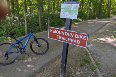 A mountain bike parked beside a sign directing to the Mountain Bike Trailhead at Maybury State Park, surrounded by lush green trees and a paved pathway. Maybury mountain bike trail.