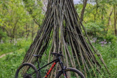 A black mountain bike with red accents resting on a grassy area near a tall, teepee-like structure made of sticks. The background features lush greenery and trees under a cloudy sky. Creekside Trails mountain bike trail.