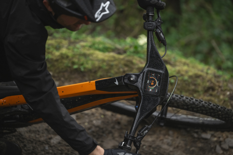 A close-up view of an electric mountain bike with a sleek orange and black frame, showcasing the digital display on the bike's top tube. A rider, dressed in dark outdoor gear and gloves, is adjusting the bike, with rain droplets visible on the frame, set against a green, natural backdrop.