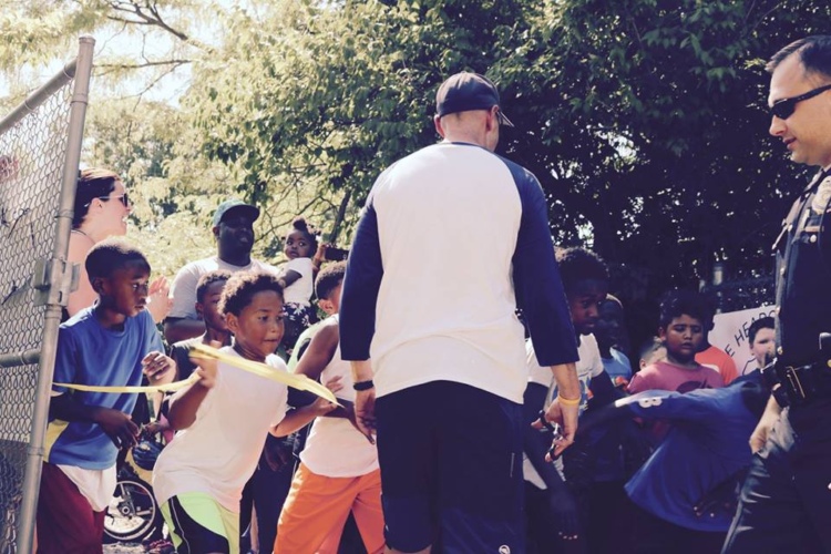 A diverse group of children excitedly gather around an adult and a police officer, with some holding a yellow ribbon. The scene takes place in a park setting, surrounded by trees. Others are watching, clapping, and smiling, creating a lively and engaging atmosphere.