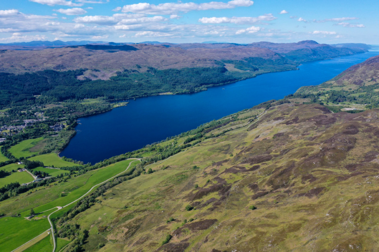 Aerial view of a serene landscape featuring a blue lake surrounded by lush green forests and rolling hills under a partly cloudy sky. The scene includes winding roads and patches of meadowland, showcasing the natural beauty of the area.
