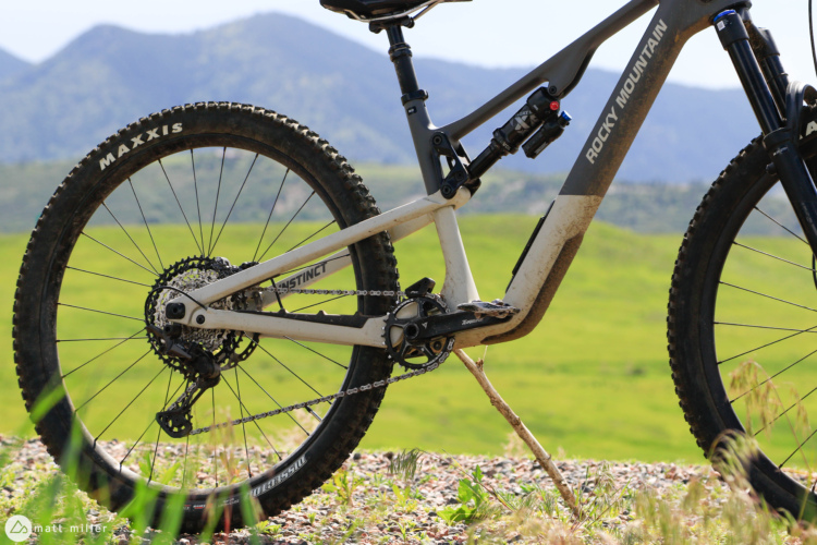 Close-up of a Rocky Mountain Instinct mountain bike, showcasing the rear wheel and drivetrain. The bike features Maxxis tires and a visible chain and gearing system. The background features green fields and mountains, indicating an outdoor setting. The image captures the bike's design and ruggedness, highlighting its suitability for trail riding.