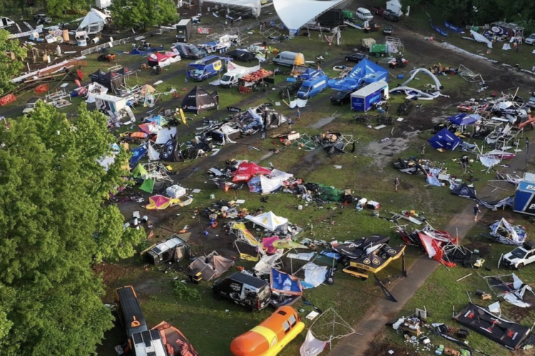 Aerial view of a festival or event area showing significant damage and debris. Tattered tents and canopies are scattered across the muddy ground, with vehicles and equipment partially visible among the wreckage. Trees can be seen in the background, and the scene conveys a sense of chaos and disorder.