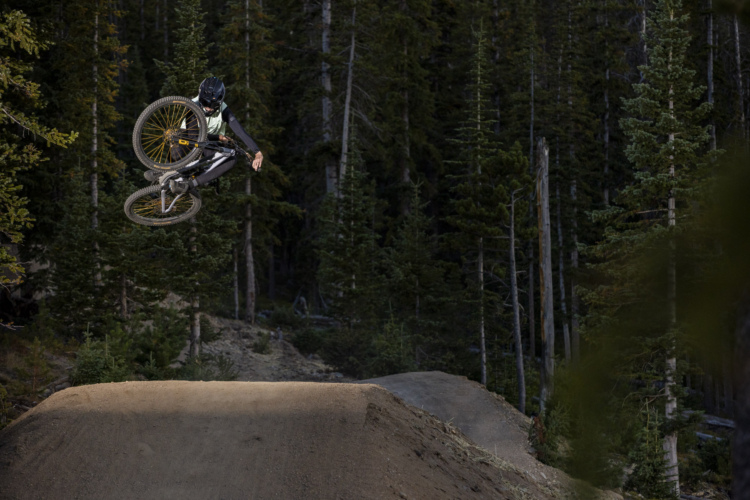 A mountain biker performing a stunt mid-air over a dirt jump, surrounded by a dense forest of pine trees. The rider is wearing a helmet and protective gear, showcasing an impressive leap with the bike’s wheels elevated above the ground.