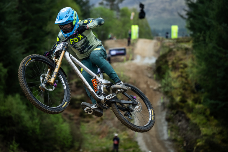 A mountain biker performs a mid-air jump over a dirt track, wearing a blue helmet and a green jersey with sponsor logos. The background features greenery and a blurred track, showcasing a competitive biking environment.