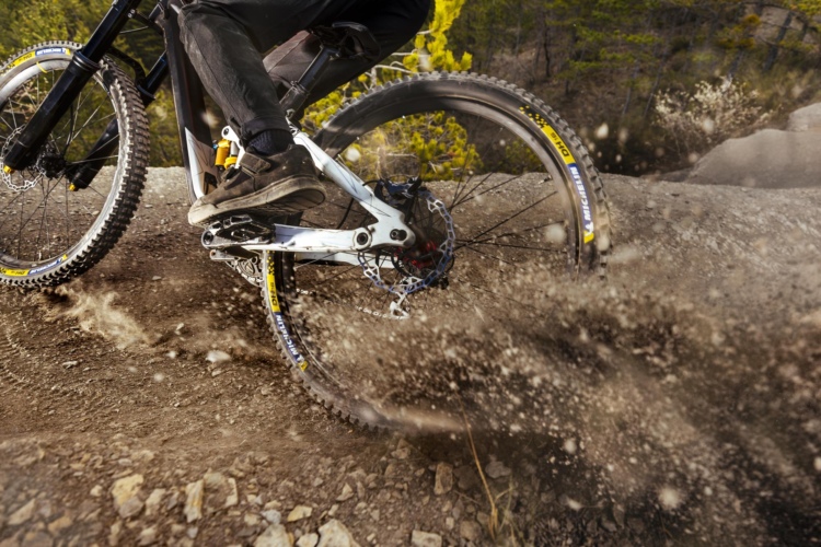 A close-up view of a mountain bike's rear wheel kicking up dirt and dust while navigating a rugged trail. The cyclist’s foot is visible on the pedal, showcasing biking shoes and the bike's details, including the drivetrain and rear suspension. Green trees are faintly visible in the background, suggesting an outdoor setting.