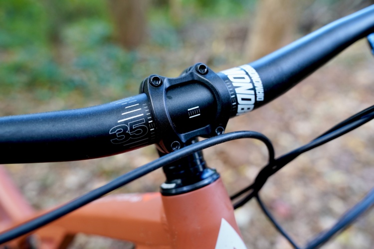 Close-up view of a mountain bike's handlebar and stem, featuring a black handlebar with measurement markings and a sturdy stem with a modern design. The background is blurred with hints of greenery, suggesting an outdoor setting.