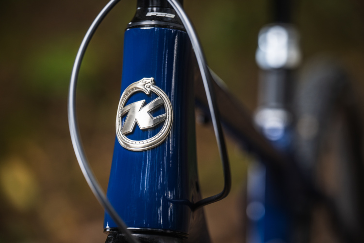 Close-up of a bicycle's head tube featuring a metallic emblem with a stylized "K" on a glossy blue finish. The image shows the bicycle's cable routing and part of the fork in a natural, softly blurred background.