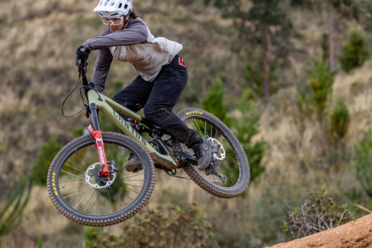 A mountain biker wearing a helmet and protective gear performs a jump on a dirt trail, showcasing skill and control in mid-air. The background features grassy hills and scattered trees, emphasizing an outdoor adventure setting.
