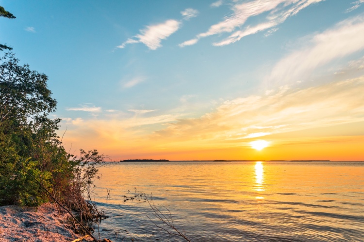 A scenic view of a tranquil lake at sunset, with the sun reflecting on the water. The sky is filled with soft pastel colors, including shades of orange, pink, and blue, while silhouetted trees frame the left side of the image. The calm water ripples gently, mirroring the beautiful sky.