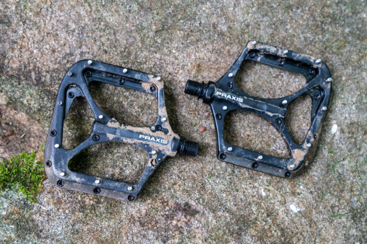 Close-up of a pair of black Praxis bicycle pedals resting on a textured stone surface, with visible dirt and mud indicating outdoor use.