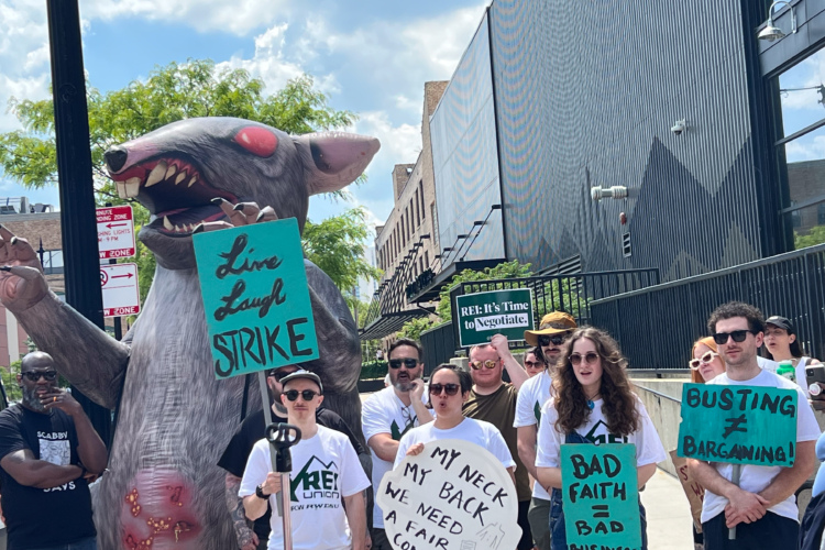 A group of protestors stands in front of a building, holding signs with messages related to a strike, including "Live Laugh Strike" and "REI: It's Time to Negotiate." A large inflatable rat is prominently featured among the crowd, symbolizing labor disputes. The protestors are dressed in a mix of white t-shirts and casual wear, some wearing sunglasses, while demonstrating solidarity for their cause under a partly cloudy sky.