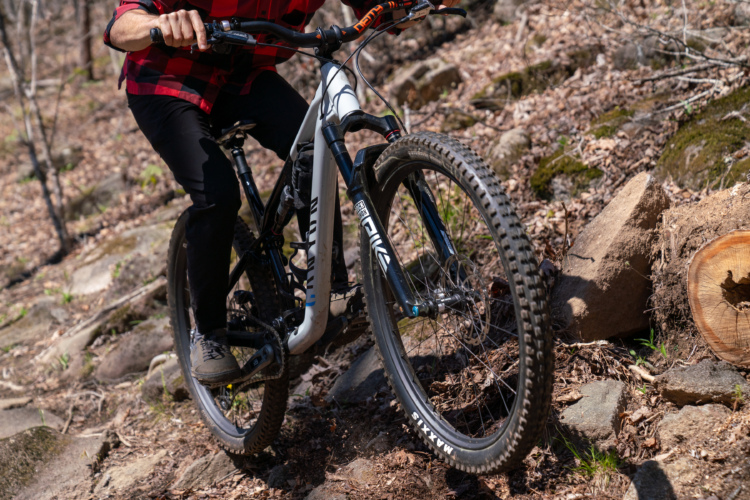 A person riding a mountain bike on a rocky trail in a wooded area, wearing a red plaid shirt and black pants. The bike features thick tires and a durable frame, navigating through a landscape with scattered rocks and fallen leaves.