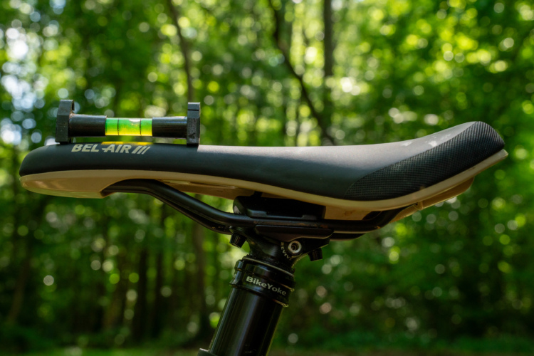 Close-up of a bicycle saddle labeled "BEL-AIR III," mounted on a black seat post with a visible leveling tool on top. The background features a blurred, lush green forest setting, indicating an outdoor cycling environment.