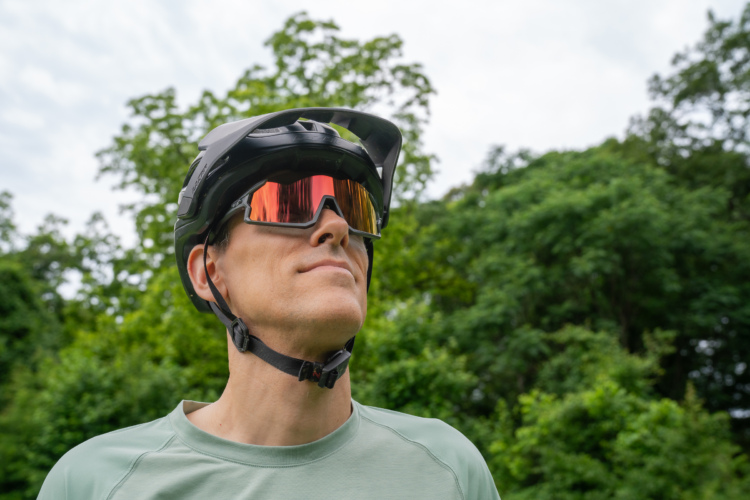 A person wearing a black cycling helmet and reflective sunglasses, looking up against a backdrop of trees and cloudy skies. The individual is dressed in a light green athletic shirt.