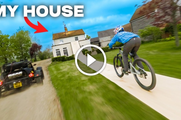 A cyclist in a blue jacket rides on a wooden path through a grassy area, with a house visible in the background. A vehicle can be seen approaching from the left. The image has an overlay indicating "MY HOUSE" with an arrow pointing toward the house.