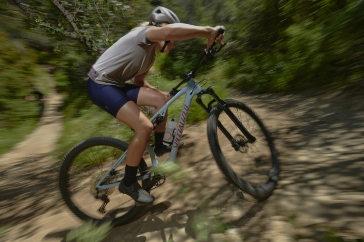 A cyclist riding a mountain bike on a dirt trail, leaning forward with one arm on the handlebars and the other braced against the bike. The background is blurred, suggesting high speed, while greenery surrounds the trail. The rider is wearing a helmet, a short-sleeved shirt, and cycling shorts.