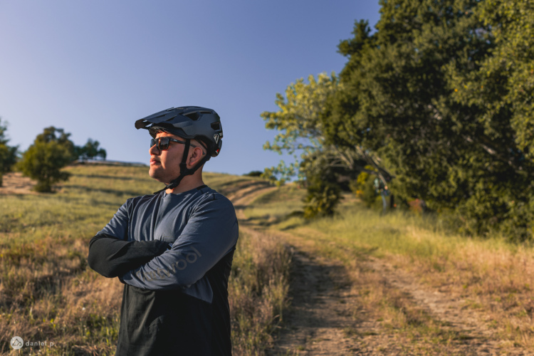 A person wearing a 7iDP Project.21 mountain bike helmet and sunglasses stands confidently with arms crossed on a dirt path surrounded by greenery and rolling hills, under a clear blue sky.