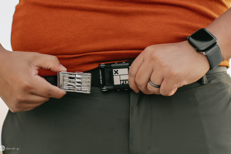 A person adjusting their belt, wearing an orange shirt and gray pants, with a close-up view of a belt buckle that has various mechanical features. The individual also wears a smartwatch on their wrist.