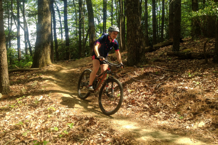 A person riding a mountain bike along a dirt trail in a forested area, surrounded by tall trees and scattered autumn leaves. The cyclist is wearing a helmet and athletic clothing, navigating a curve in the trail.