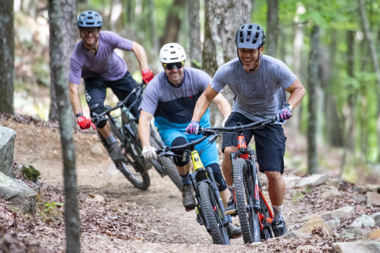 Three mountain bikers riding on a forest trail, surrounded by trees and rocks. The cyclists are wearing helmets and casual sportswear, and they appear to be enjoying an energetic ride through the greenery. One rider is leaning into a turn, while the others follow closely behind. The ground is a mix of dirt and scattered leaves, typical of a wooded biking path.