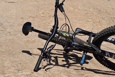A mountain bike resting on a rocky, uneven terrain under a clear blue sky. The landscape features a distant path that winds up the rocky surface, surrounded by sparse vegetation. Slickrock mountain bike trail.