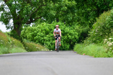 A cyclist wearing a colorful jersey and helmet rides along a narrow country road surrounded by lush green trees and shrubs.