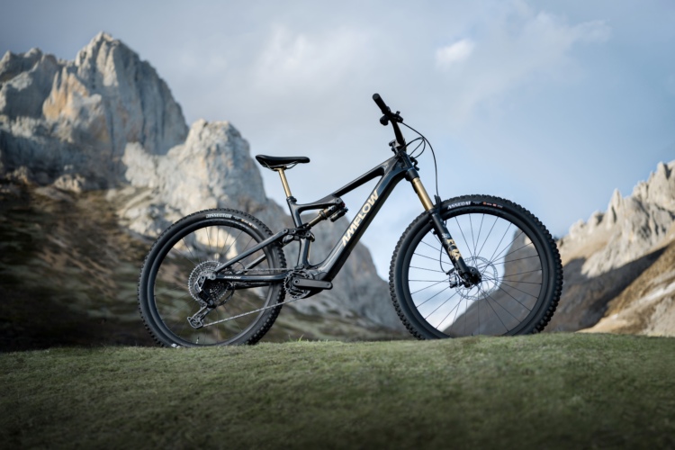 A sleek black mountain bike positioned on a green grassy slope, with rugged mountainous terrain in the background under a cloudy sky. The bike features wide tires and a modern design, emphasizing its suitability for off-road adventures.