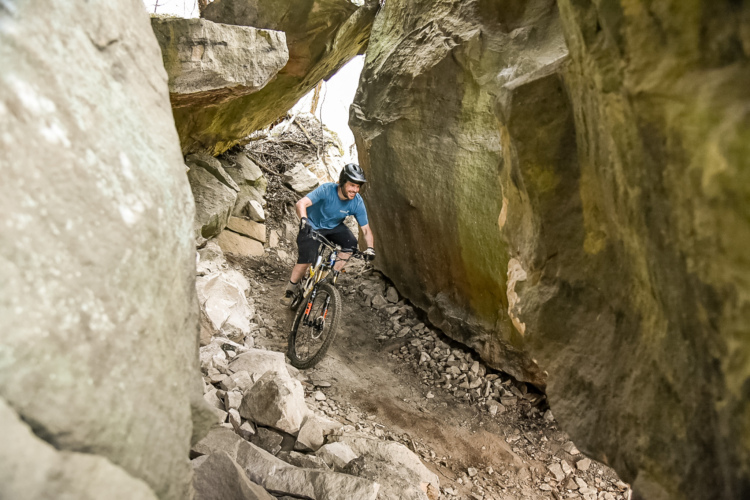 A mountain biker navigating a narrow trail between large rock formations, surrounded by rough terrain and some greenery. The biker is wearing a helmet and a blue shirt, focused on maneuvering through the rocky landscape.