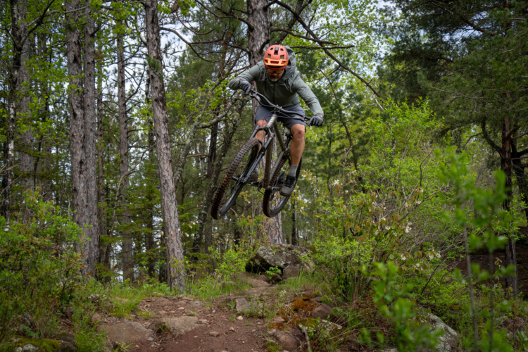 A mountain biker wearing an orange helmet and a green long-sleeved shirt jumps over a rocky path in a forested area, surrounded by trees and greenery. The bike’s wheels are off the ground, showcasing an action shot of the rider mid-air.