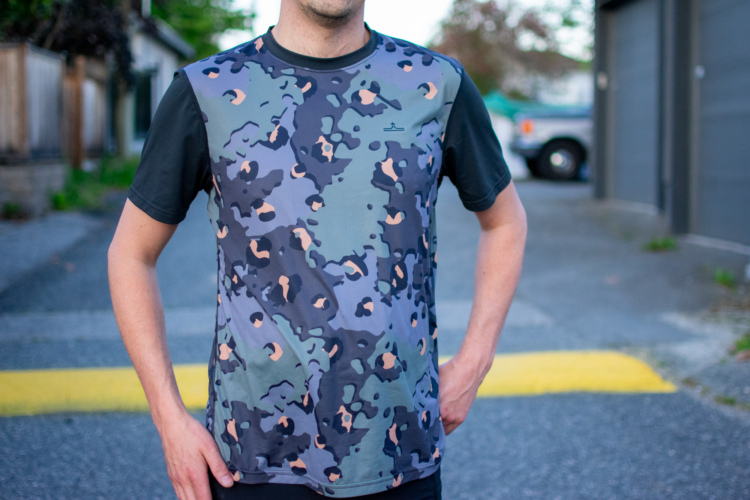 A person standing outdoors wearing a short-sleeve, camouflage-patterned athletic shirt in shades of green, gray, and peach. They are posed with one hand on their hip, set against a blurred urban background.